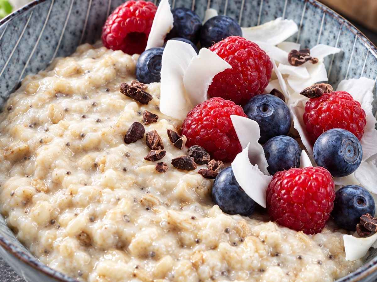 Nahaufnahme von cremigem Kokos-Porridge mit Himbeeren, rosa Beerenschlieren und knusprigen Kokoschips